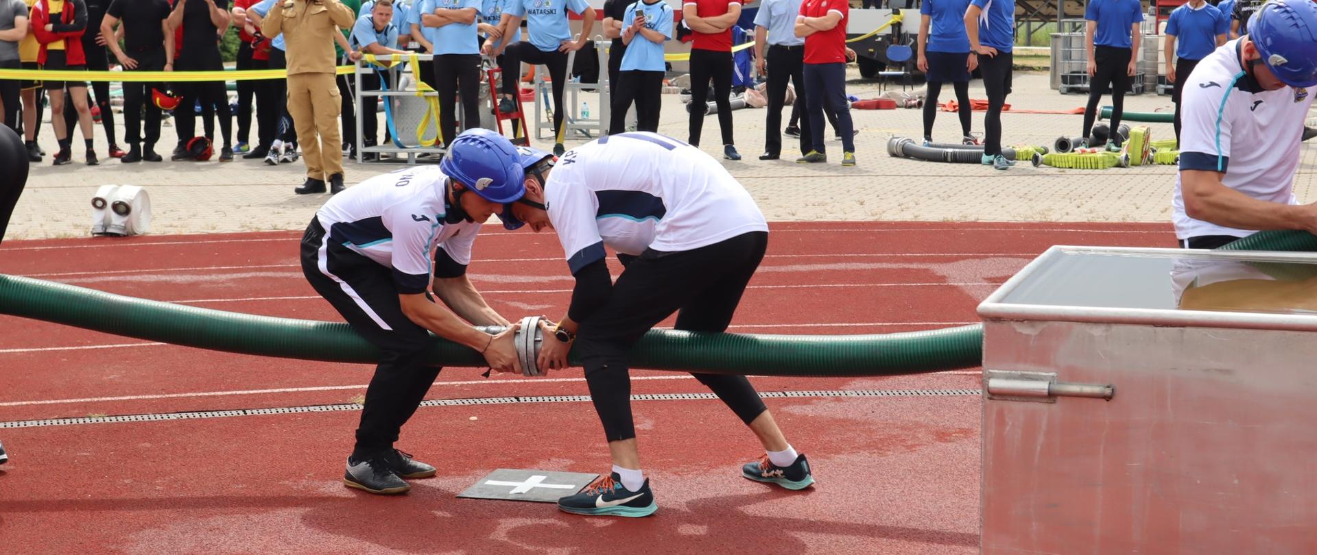 Widok na zawodników strażaków, którzy sprawiają linię wężową ssawną. W tle obiekt ośrodka oraz kilkadziesiąt osób obserwujących.