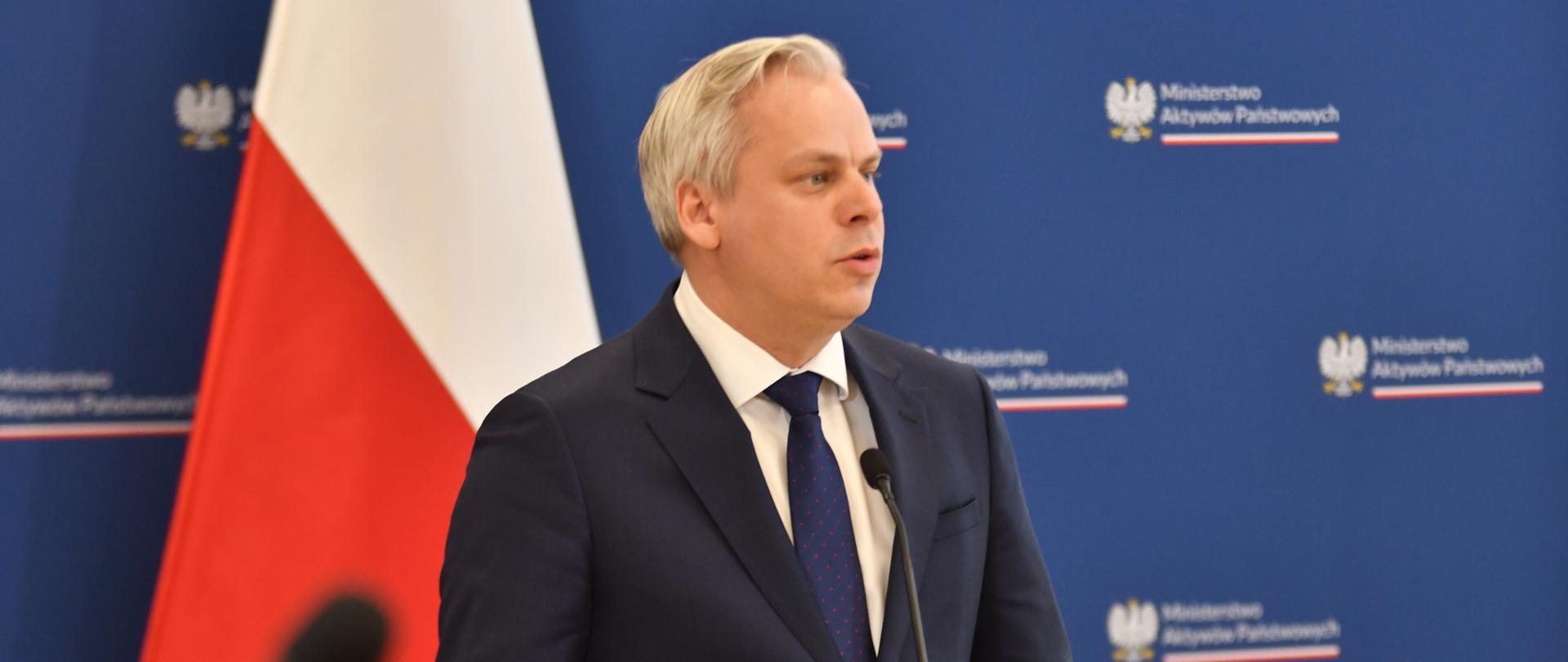 Wiceminister Karol Rabenda na briefingu prasowym, przemawia przy mównicy. W tle polska flaga oraz ścianka z logotypem MAP