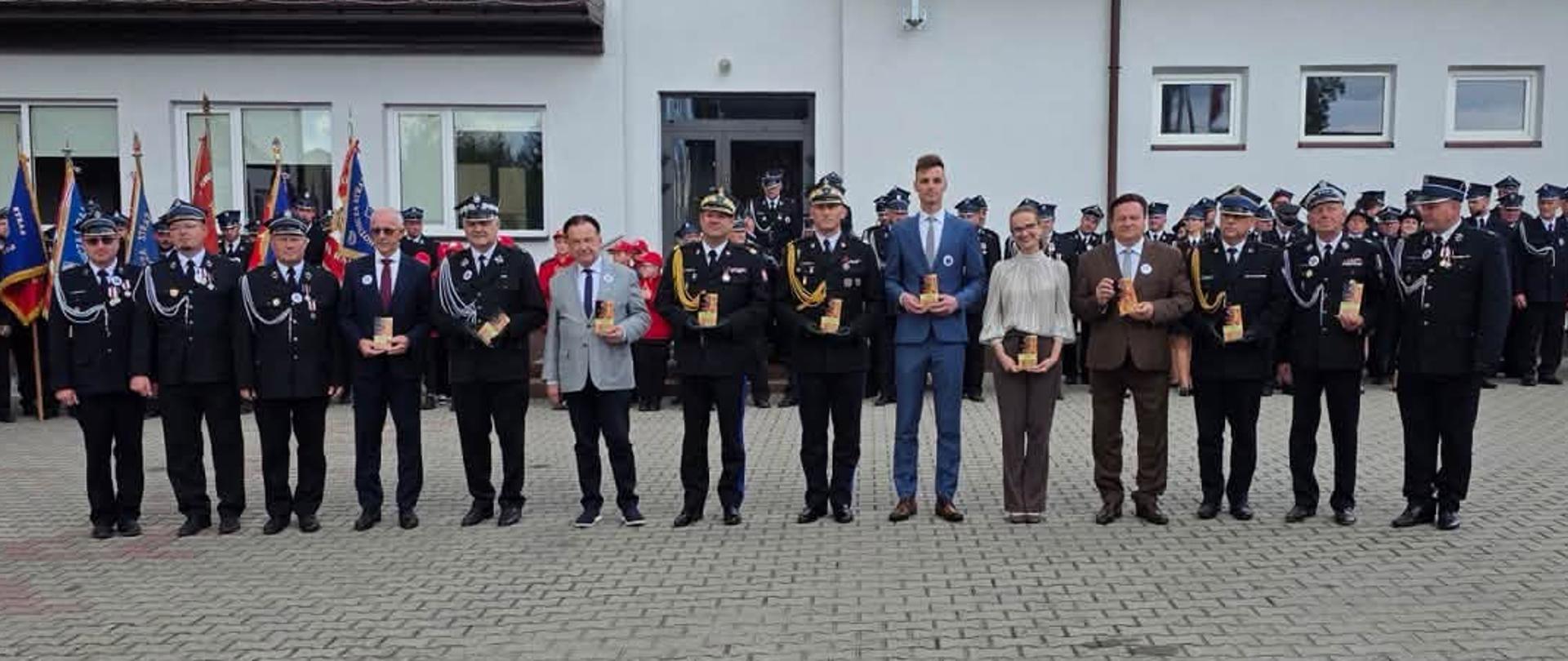 Poziome zdjęcie przedstawia rząd kilkunastu strażaków oraz cywilnych dostojników stojących wzdłuż brukowanego placu po zakończeniu ceremonii odznaczeń. Druhowie ubrani są w ciemnogranatowe mundury galowe z czapkami rogatywkami. Na ich piersiach błyszczą nowo wręczone medale (m.in. Złoty Znak Związku, Medale za Zasługi dla Pożarnictwa, Odznaki Świętego Floriana), a w dłoniach trzymają czerwone etui oraz okolicznościowe dyplomy. W tle po prawej stronie widoczni są zgromadzeni mieszkańcy oraz fragment białego budynku strażnicy OSP. Pogoda jest jasna z lekkim zachmurzeniem. Ze zdjęcia bije ogólna duma, radość, satysfakcja z dobrze wypełnionej misji oraz satysfakcja z zasłużonych wyróżnień.