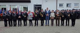 Poziome zdjęcie przedstawia rząd kilkunastu strażaków oraz cywilnych dostojników stojących wzdłuż brukowanego placu po zakończeniu ceremonii odznaczeń. Druhowie ubrani są w ciemnogranatowe mundury galowe z czapkami rogatywkami. Na ich piersiach błyszczą nowo wręczone medale (m.in. Złoty Znak Związku, Medale za Zasługi dla Pożarnictwa, Odznaki Świętego Floriana), a w dłoniach trzymają czerwone etui oraz okolicznościowe dyplomy. W tle po prawej stronie widoczni są zgromadzeni mieszkańcy oraz fragment białego budynku strażnicy OSP. Pogoda jest jasna z lekkim zachmurzeniem. Ze zdjęcia bije ogólna duma, radość, satysfakcja z dobrze wypełnionej misji oraz satysfakcja z zasłużonych wyróżnień.