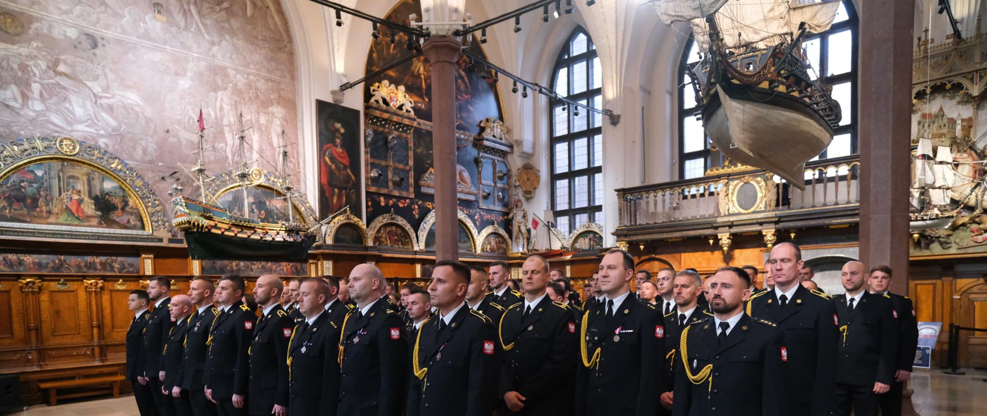 Strażacy stoją w szyku w Dworze Artusa podczas uroczystych obchodów Dnia Niepodległości.