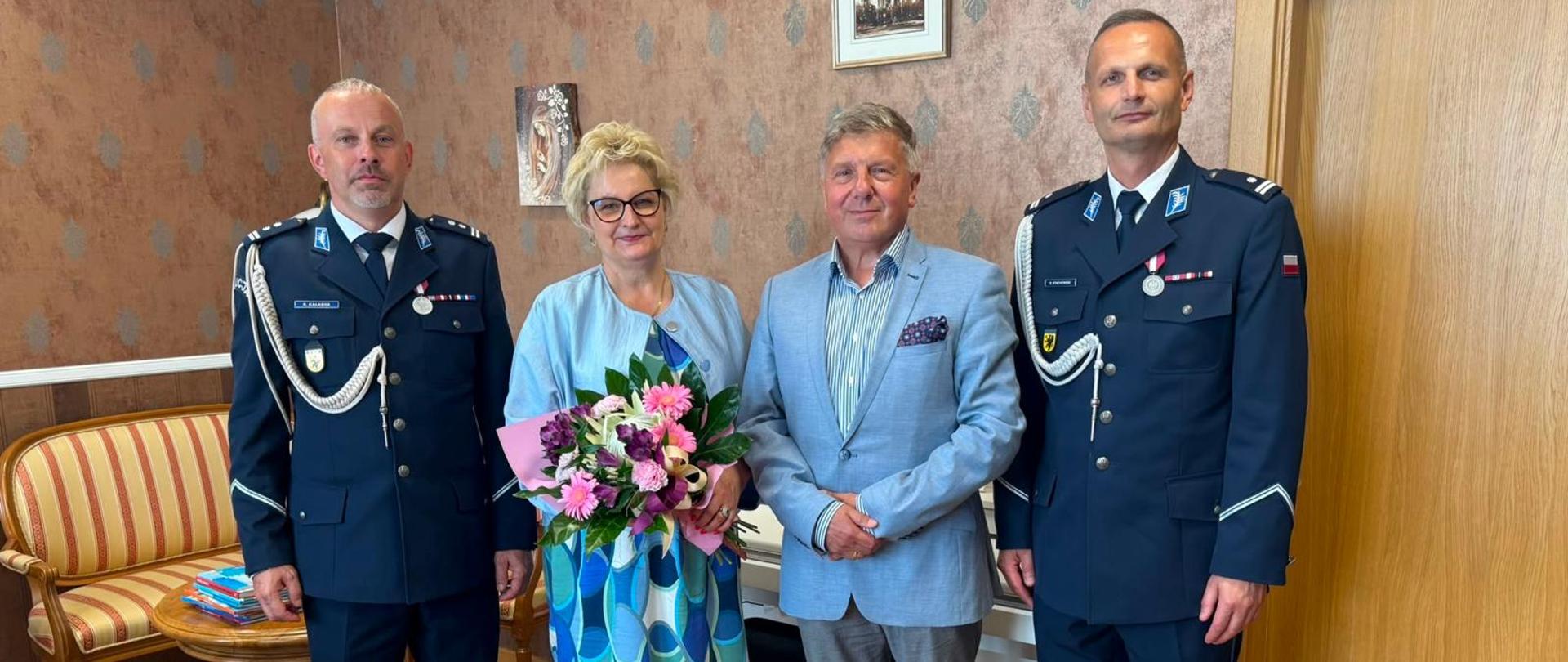 Zdjęcie wykonane podczas obchodów Święta Policji. Po lewej stronie mężczyzna w mundurze policyjnym. Dalej do prawej strony kobieta blondynka w okularach, błękitnej sukni w koła i marynarce, która trzyma bukiet kwiatów. Obok niej mężczyzna w szarych spodniach i błękitnej marynarce. Na samym końcu po prawej stron ie mężczyzna w mundurze policyjnym. Tło obrazka w odcieniach brązu.