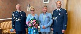 Zdjęcie wykonane podczas obchodów Święta Policji. Po lewej stronie mężczyzna w mundurze policyjnym. Dalej do prawej strony kobieta blondynka w okularach, błękitnej sukni w koła i marynarce, która trzyma bukiet kwiatów. Obok niej mężczyzna w szarych spodniach i błękitnej marynarce. Na samym końcu po prawej stron ie mężczyzna w mundurze policyjnym. Tło obrazka w odcieniach brązu.
