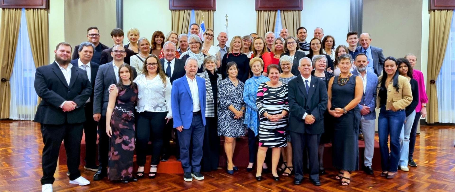 Na zdjęciu widać dużą, elegancko ubraną grupę ludzi pozujących do wspólnej fotografii w jasnej, reprezentacyjnej sali.
Opis szczegółowy:
Grupa liczy około czterdziestu kilku osób, stojących w kilku rzędach. Wszyscy patrzą w stronę aparatu i uśmiechają się. Wiek uczestników jest zróżnicowany. Wiele osób ma na sobie ubrania wizytowe: garnitury, marynarki, koszule, sukienki koktajlowe, garsonki. Atmosfera jest uroczysta.