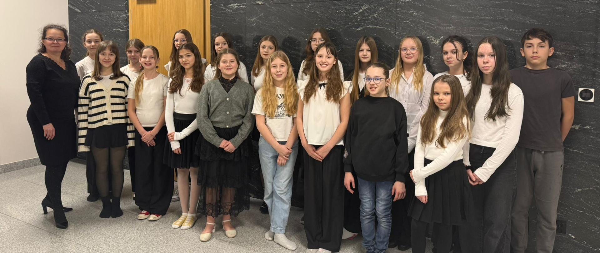 Grupa uczniów szkolnego chóru wraz z nauczycielką stoi w holu przed wejściem do sali koncertowej. Na ścianie za nimi widoczny jest napis „SALA KONCERTOWA” oraz oznaczenie „WEJŚCIE 1”. Młodzież ubrana jest głównie w biało-czarne stroje galowe. Uczniowie ustawieni są w kilku rzędach i pozują do wspólnego zdjęcia.