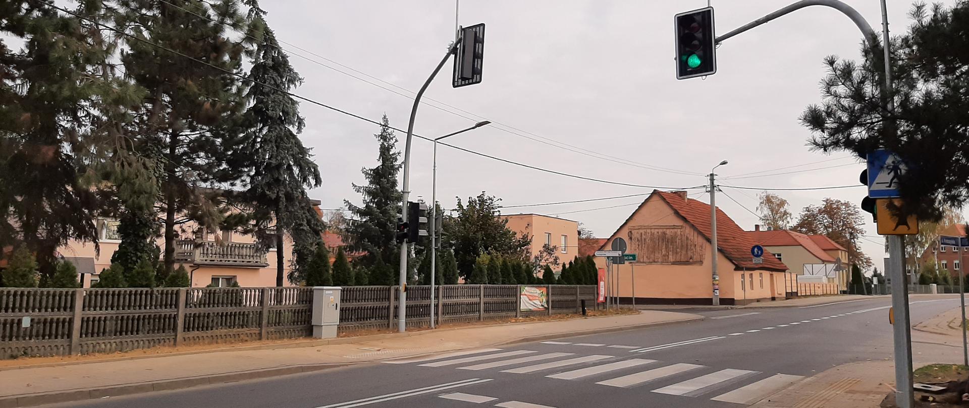 centralnie widoczne przejście dla pieszych na jezdni, na chodnikach po obu stronach przejścia widoczna nowa sygnalizacja świetlna. Droga przebiega od lewej strony zdjęcia do prawej. Po lewej stronie w tle widoczne zabudowania mieszkalnie, domy jednorodzinne.