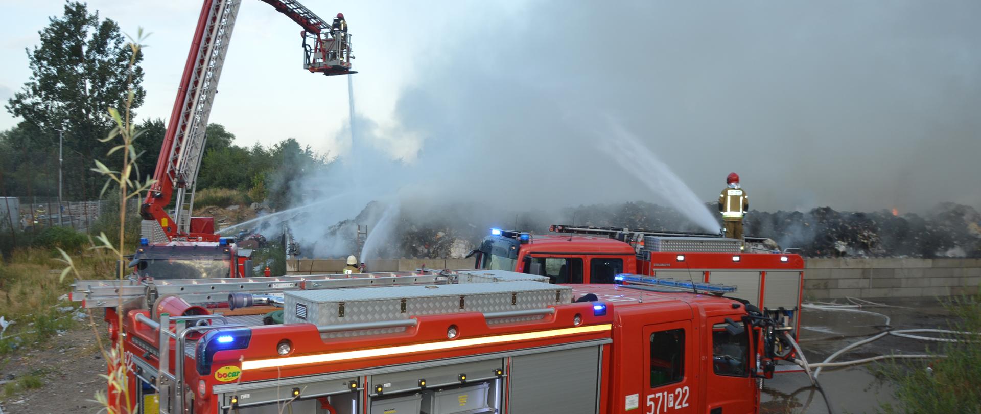 Pożar na składowisku odpadów. Plac składowy, a na nim trzy wozy strażackie, w tym jeden z podnośnikiem hydraulicznym. Prowadzone są działania gaśnicze. Jeden ze strażaków podaje w wodę z działka, stojąc na dachu samochodu, drugi - z ziemi, a trzeci z kosza podnośnika. Występuje silne zadymienie, widoczne są płomienie.