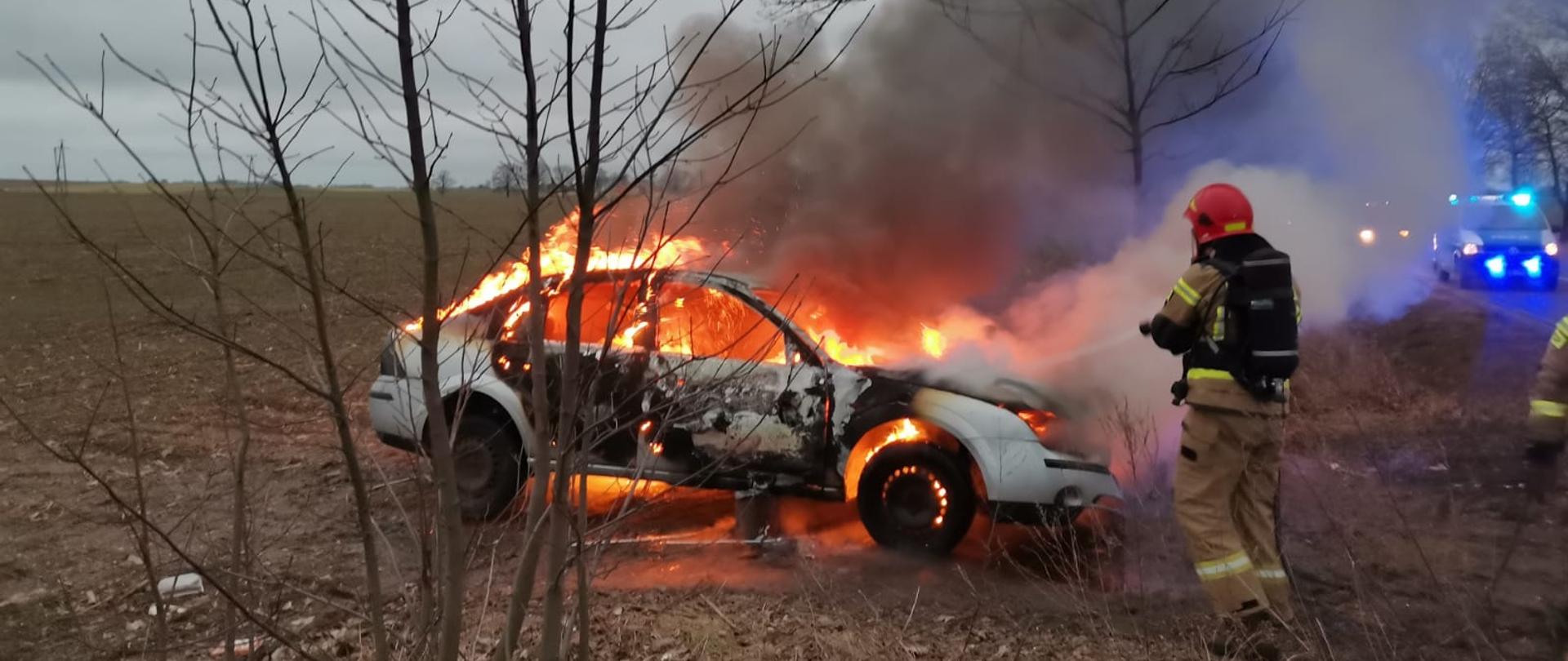 Fotografia przedstawia widok płonącego samochodu osobowego marki Ford Focus, ogniem objęty jest cały pojazd, widoczne znaczne zadymienie za pojazdem. Samochód stoi na poboczu drogi, w tle widoczne zaorane pole. Ujęcie wykonane z Drogi w kierunku pola. Przy pojeździe stoi strażak z prądownicą, gasi płonący pojazd. Strażak w ubraniu specjalnym, z założonym aparatem ochrony dróg oddechowych oraz w hełmie z opuszczoną przyłbicą, stoi częściowo tyłem do fotografującego. po prawej stronie zdjęcia na szosie widoczny radiowóz blokujący ruch na jednym z pasów.