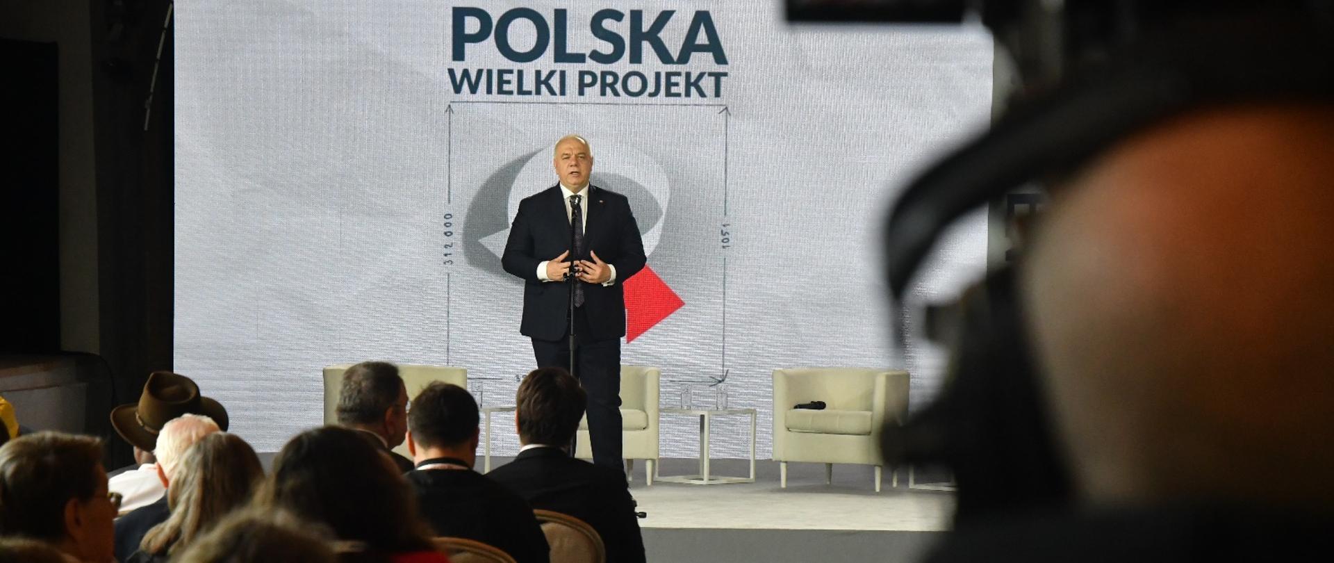 Wicepremier Jacek Sasin stoi za mikrofonem. Ujęcie zza słuchaczy. 