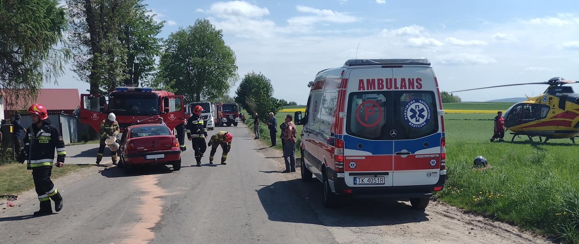 Na zdjęciu służby ratownicze (straż pożarna, pogotowie i śmigłowiec LPR) zlokalizowane na drodze powiatowej i usuwające skutki wypadku drogowego. Na jezdni widzimy uszkodzony czerwony samochód Renault. Strażacy posypują asfalt sorbentem. Po prawej stronie na terenie zielonym znajduje się śmigłowiec LPRu. 