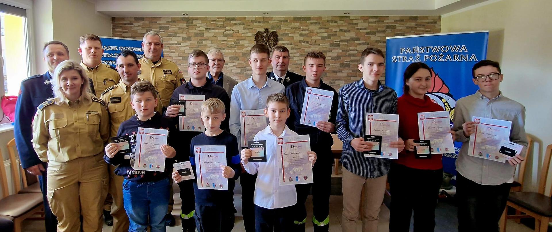 Sala sztandarowa KP PSP Tczew. Do zdjęcia ustawili się: na pierwszym planie dzieci z dyplomami oraz nagrodami - kartami podarunkowymi. Z tyłu członkowie komisji, Komendant, vice starosta