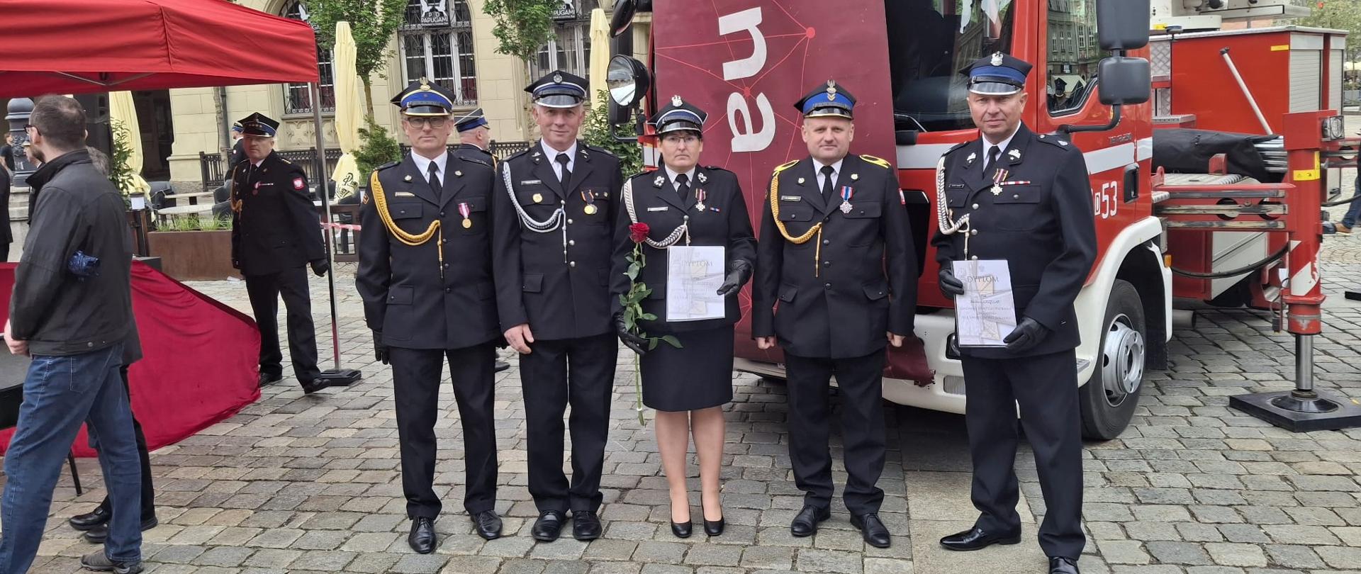 Pięcioro strażaków i jedna strażaczka w galowych mundurach pozuje do zdjęcia na rynku miejskim obok czerwonego wozu strażackiego z podnośnikiem. Dwie osoby trzymają dyplomy, a strażaczka dodatkowo czerwoną różę. W tle widać historyczne kamienice, namiot wydarzenia oraz inne osoby uczestniczące w uroczystości.
