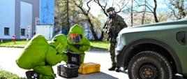 Dwóch strażaków ubranych w zielone ubrania gazoszczelne klęczą przed sprzętem. Obok stoi zielony samochód, a dalej żołnierz ubrany w moro i maskę gazoszczelną. W tle widać zabudowania zakładów i drzewa.