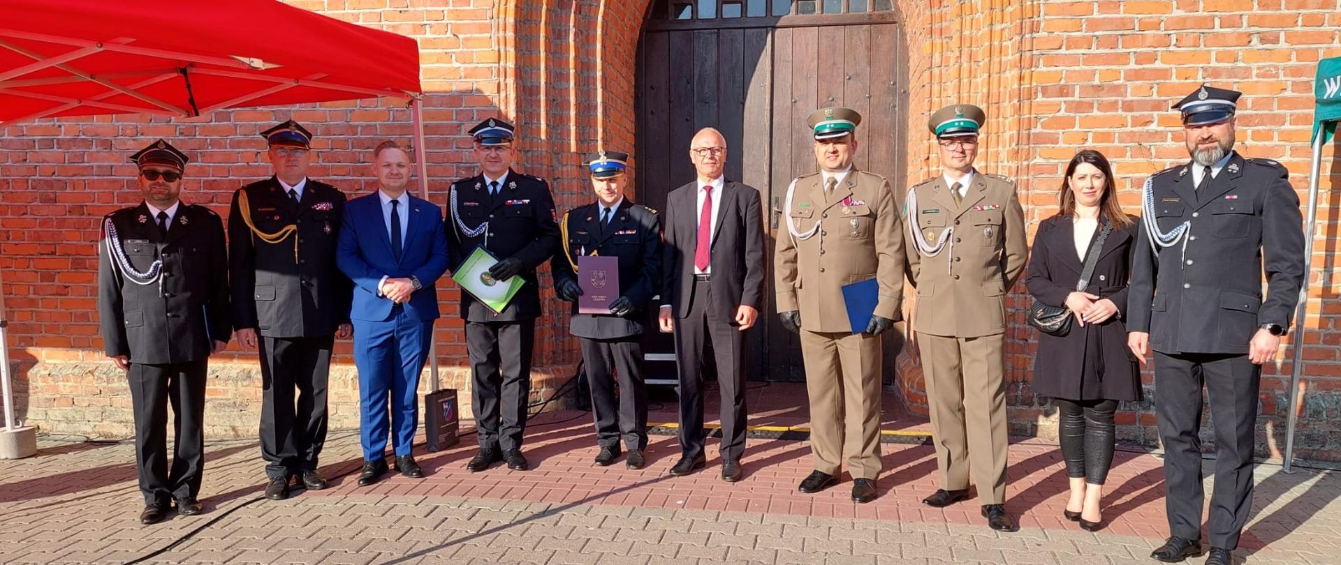 Przed obiektem kościoła z czerwonej cegły stoi grupa przedstawicieli samorządów oraz funkcjonariuszy z różnych formacji w mundurach galowych. Słonecznie.