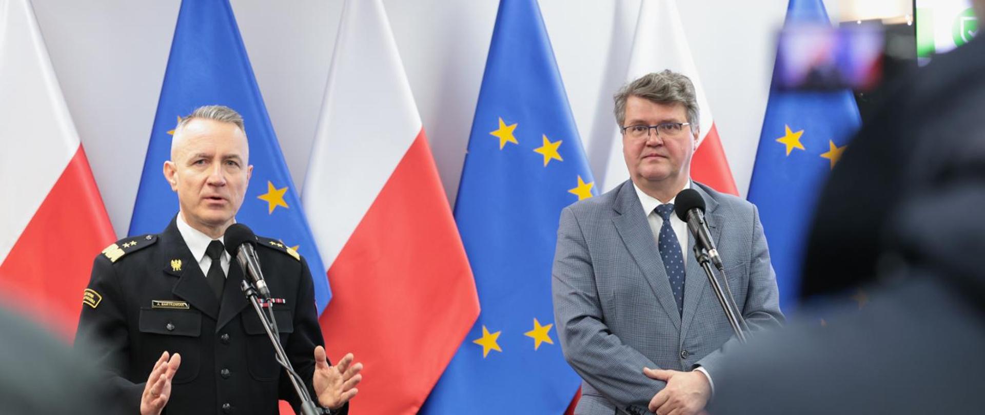 Na tle flagi Polski oraz Unii Europejskiej stoi dwóch mężczyzn. Jeden ubrany w czarny mundur wyjściowy PSP, drugi ubrany w garnitur. Obydwaj mężczyźni stoją przed mikrofonami.
