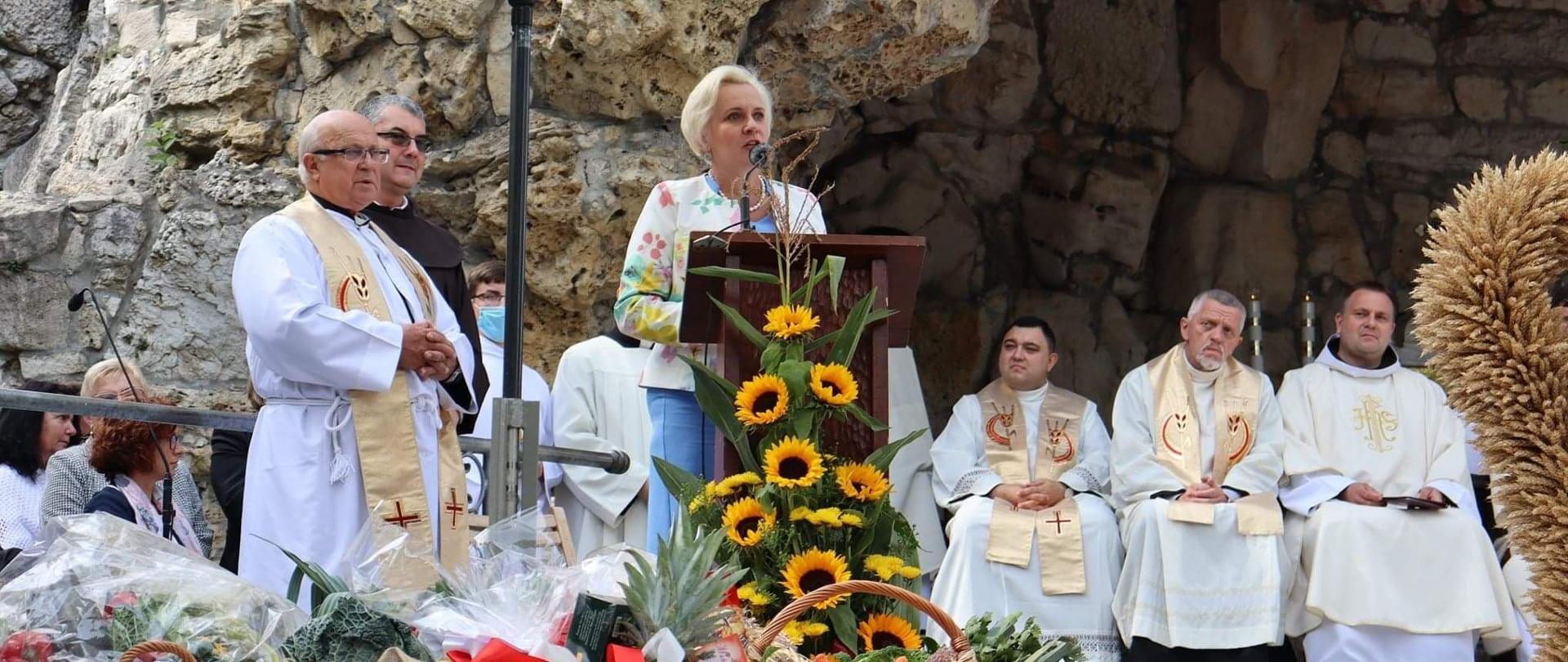 Podczas uroczystości Teresa Barańska podziękowała rolnikom, w centrum przemawiająca Teresa Barańska, przed nią kompozycja kwiatów słonecznika, po lewej i prawej stronie przysłuchujący się ludzie, z tyłu grota świętej Anny