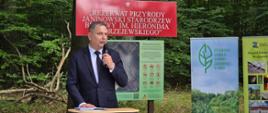 Zdjęcie przedstawia zbliżenie na mężczyznę przemawiającego do mikrofonu podczas oficjalnego wydarzenia w lesie.