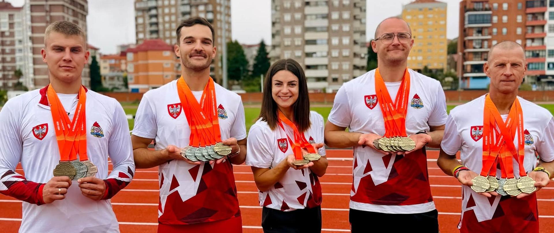 Pięciu funkcjonariuszy stoi w szeregu w miejscu rozgrywek lekkoatletycznych trzymając zdobyte medale.