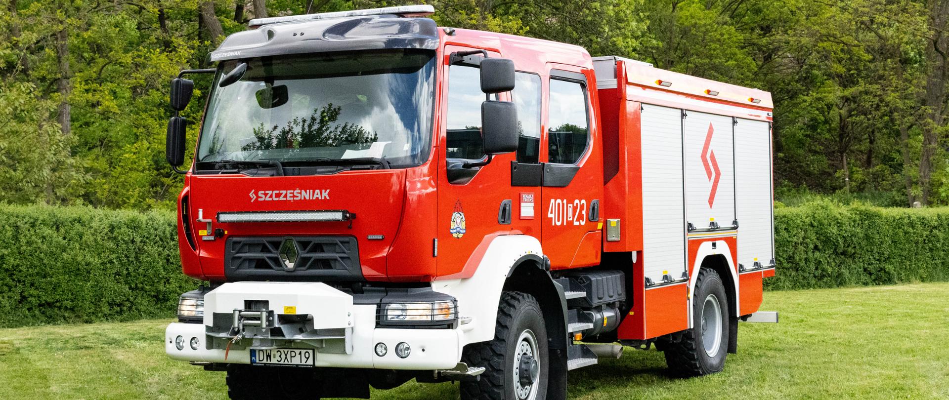 Nowoczesny, czerwony wóz strażacki Renault D15 z białym przednim zderzakiem, wyciągarką oraz zamontowaną nad grillem listwą LED (lightbar). Na boku kabiny widnieje numer „401[D]23”, a na roletach skrytek – czerwone logo Szczęśniak. Pojazd stoi na trawniku.