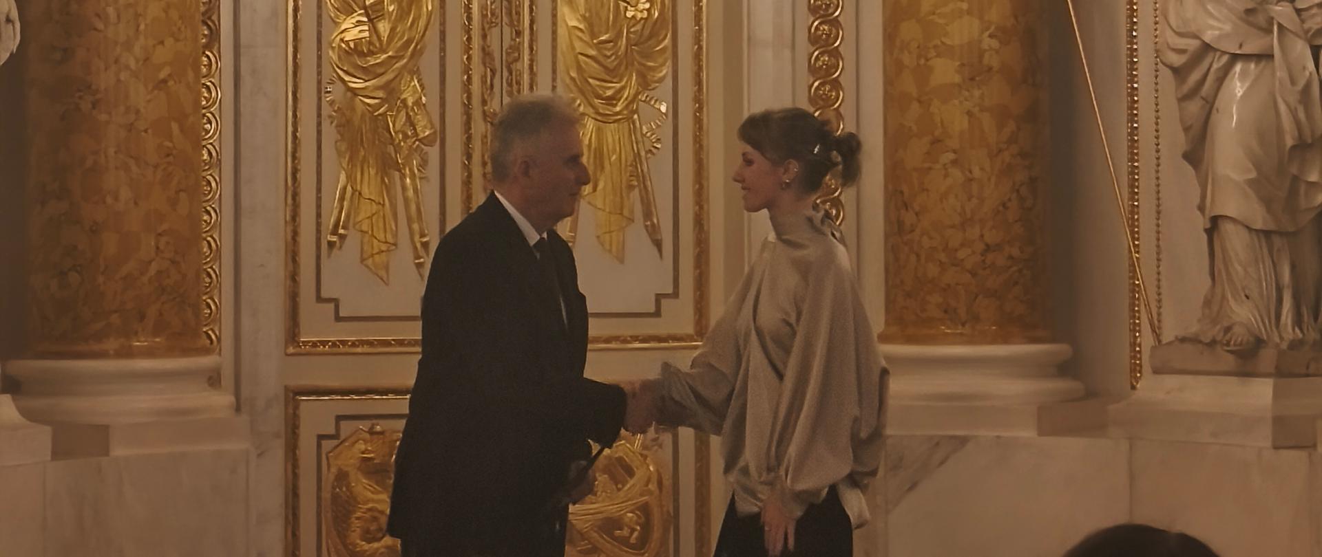 W barokowej biało-złotej sali mężczyzna gratuluje młodej dziewczynie, na pierwszym planie głowy uczestników