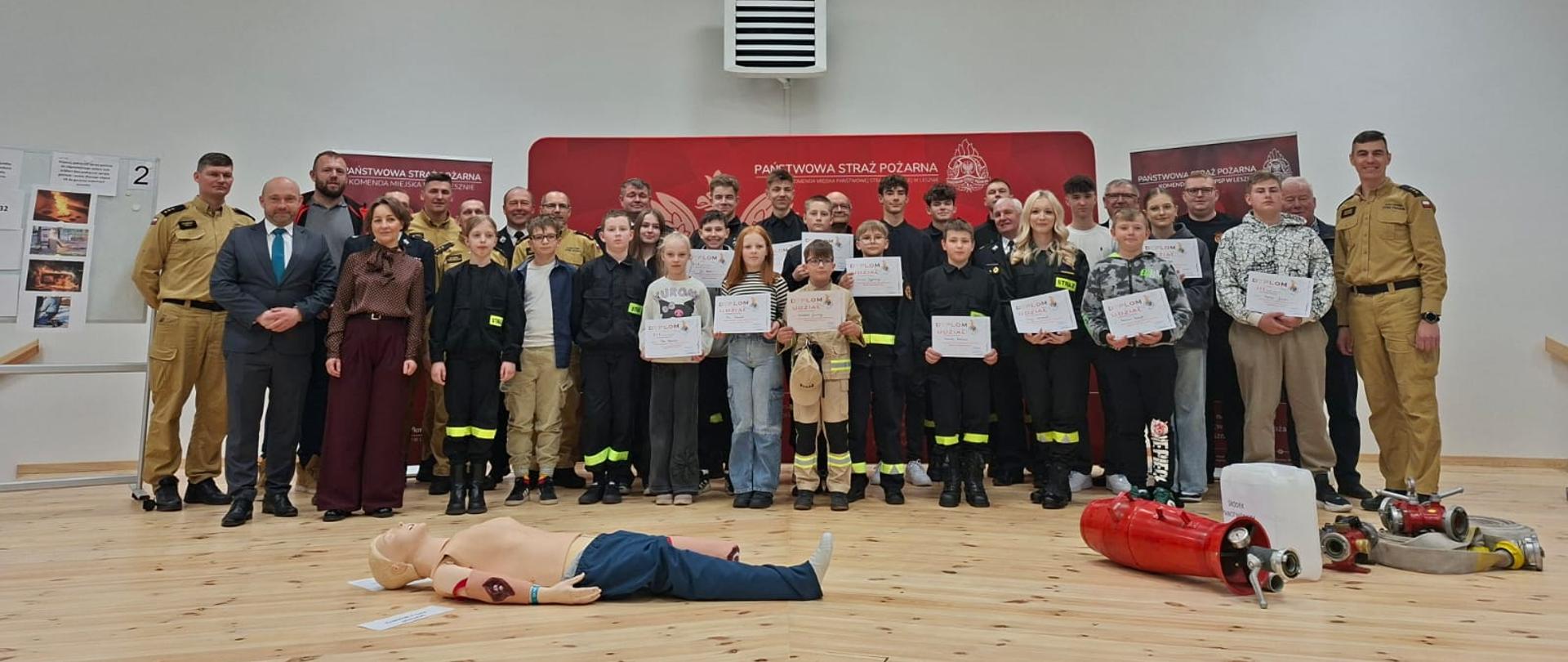 Młodzież wraz z osobami cywilnymi i strażakami stoją na scenie w trzech rzędach.