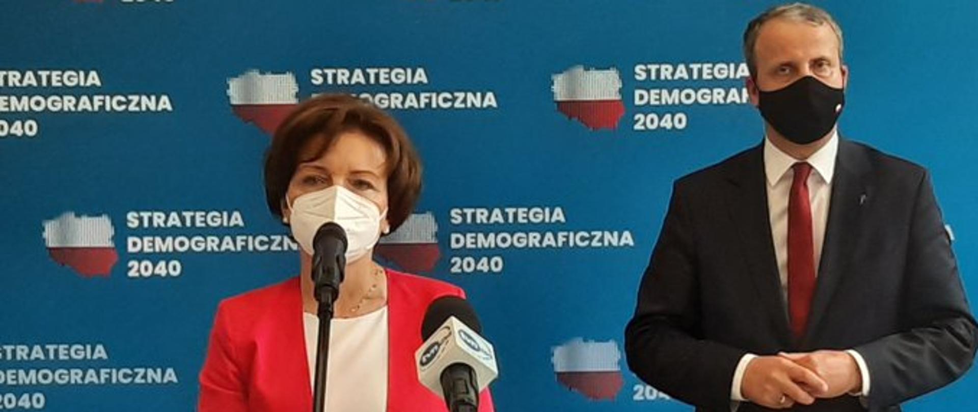 Minister Maląg wraz z wojewodą wielkopolskim stoją na tle niebieskiego baneru. Na nim napis Strategia Demograficznej 2040 obok napisu zarys geograficzny Polski wypełniony bialo-czerwonym kolorem