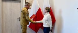 Pomorski komendant wojewódzki Państwowej Straży Pożarnej uściska dłoń funkcjonariuszowi służby cywilnej, trzymając czerwoną teczkę. Osoby stoją na tle flagi Rzeczypospolitej Polskiej.