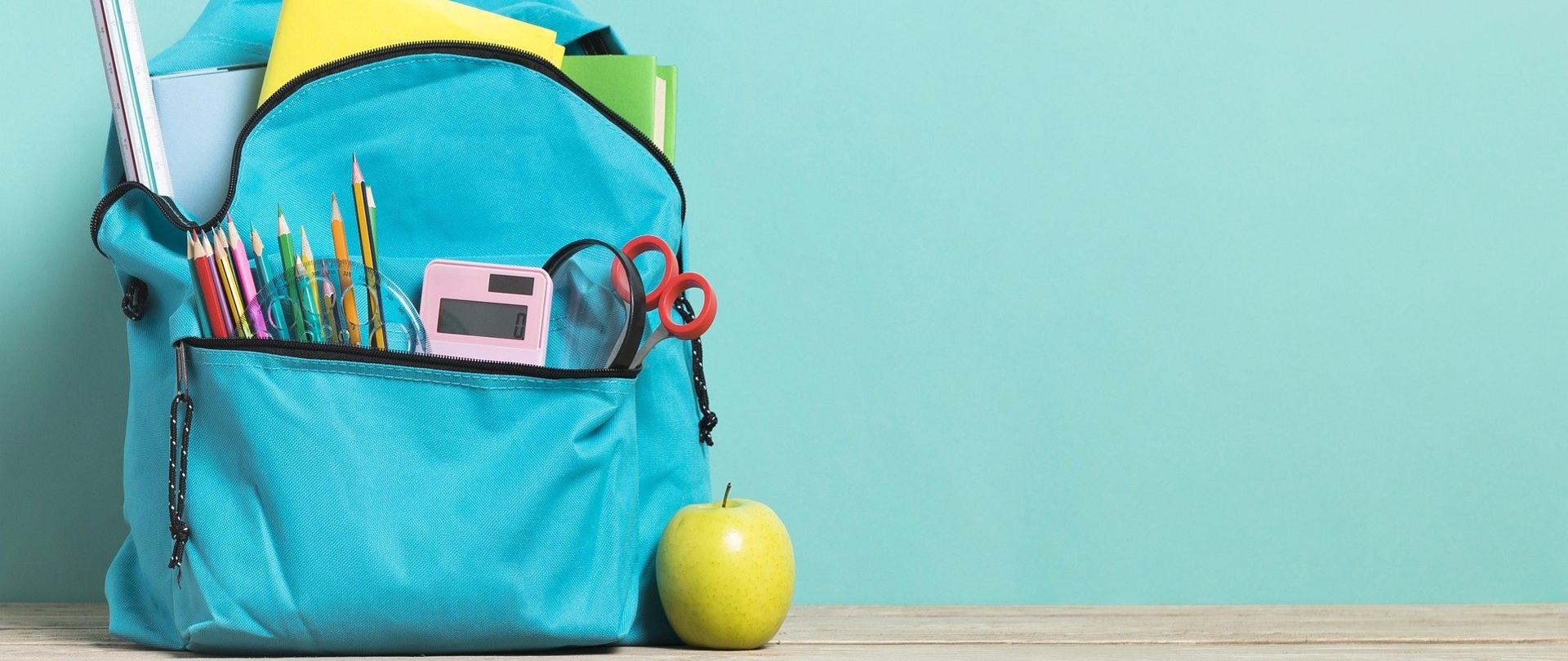 plecak szkolny stojący na podłodze pod ścianą, z którego wystają zeszyty, linijki, kredki. Przy plecaku leży zielone jabłko.