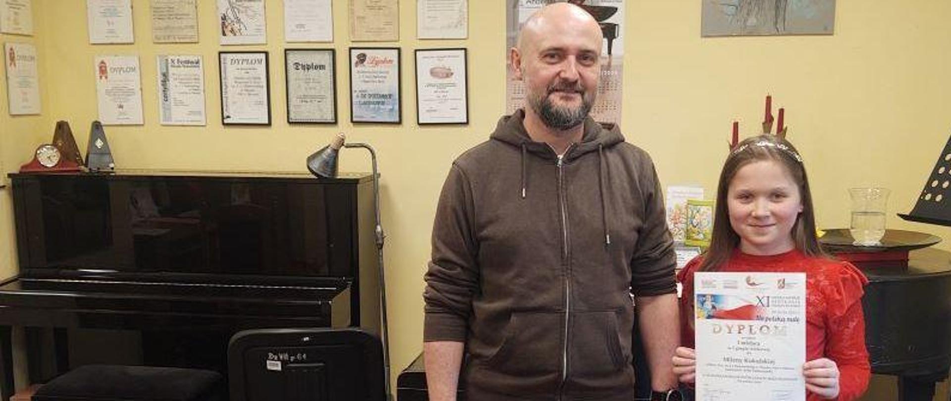 Zdjęcie kolorowe. Od lewej stoją: Artur Pastuszyński nauczyciel gry na skrzypcach, obok stoi jego uczennica Milena Kukulska ubrana w czerwoną bluzkę i czarne spodnie oraz trzyma dyplom za zajęcie I miejsca w XI Włocławskich Spotkaniach Skrzypcowych.
