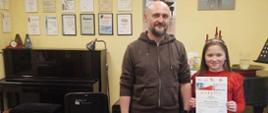 Zdjęcie kolorowe. Od lewej stoją: Artur Pastuszyński nauczyciel gry na skrzypcach, obok stoi jego uczennica Milena Kukulska ubrana w czerwoną bluzkę i czarne spodnie oraz trzyma dyplom za zajęcie I miejsca w XI Włocławskich Spotkaniach Skrzypcowych.