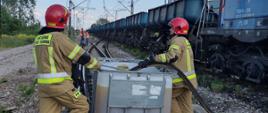 Strażacy na torowisku na tle lokomotywy z wagonami towarowymi przepompowują paliwo do mauzera
