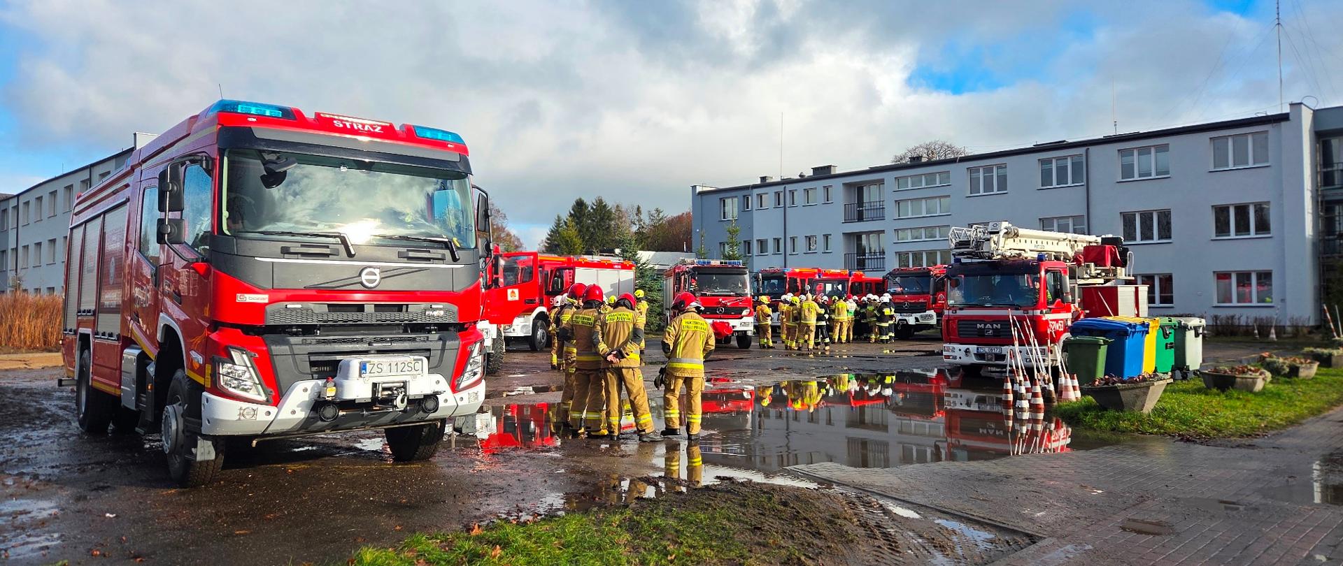  Kilku strażaków w pełnym umundurowaniu stoi obok rozstawionego czerwonego namiotu i wozu strażackiego zaparkowanego na chodniku.