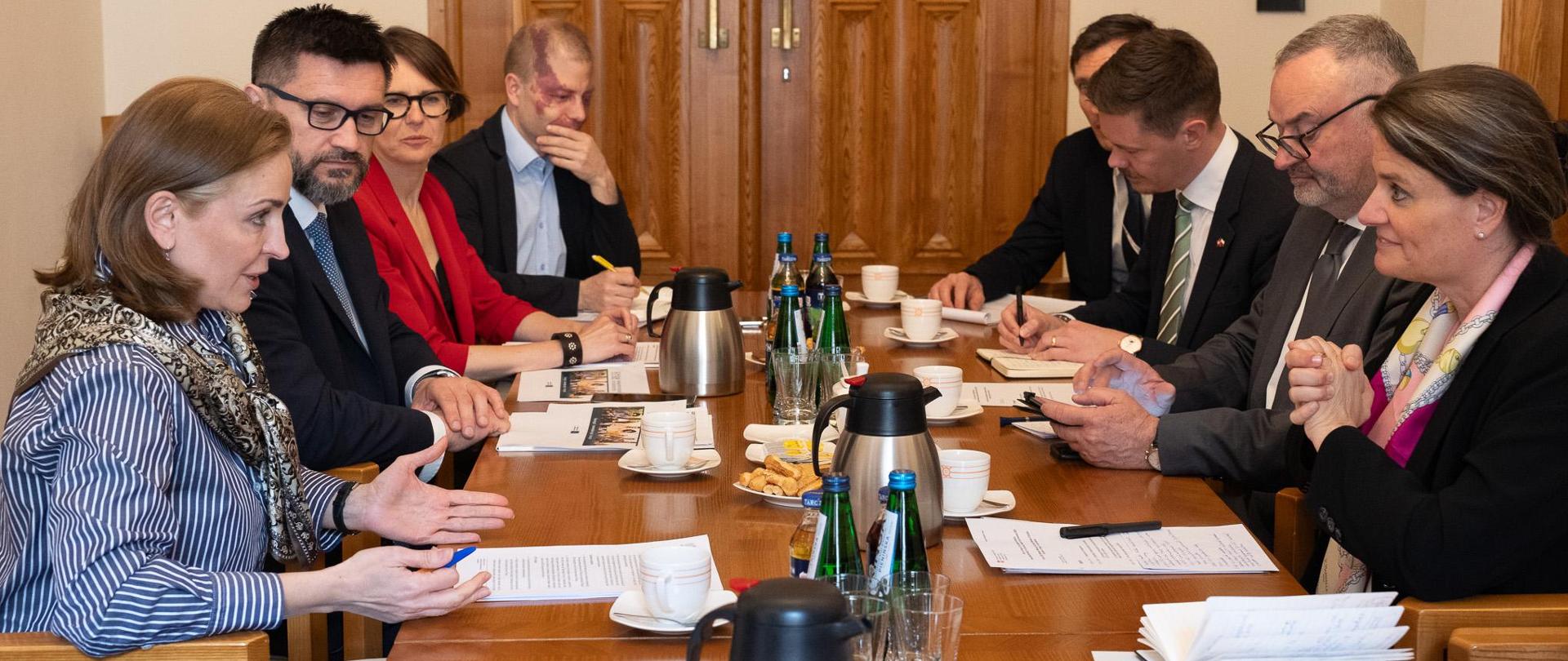 Przy stole siedzą uczestnicy spotkania. Po lewej stronie zasiada strona polska wraz z wiceminister Joanną Muchą, a po prawej strona Szwajcarska. Na stole znajdują się notatki, dokumenty, a także napoje i poczęstunek. 