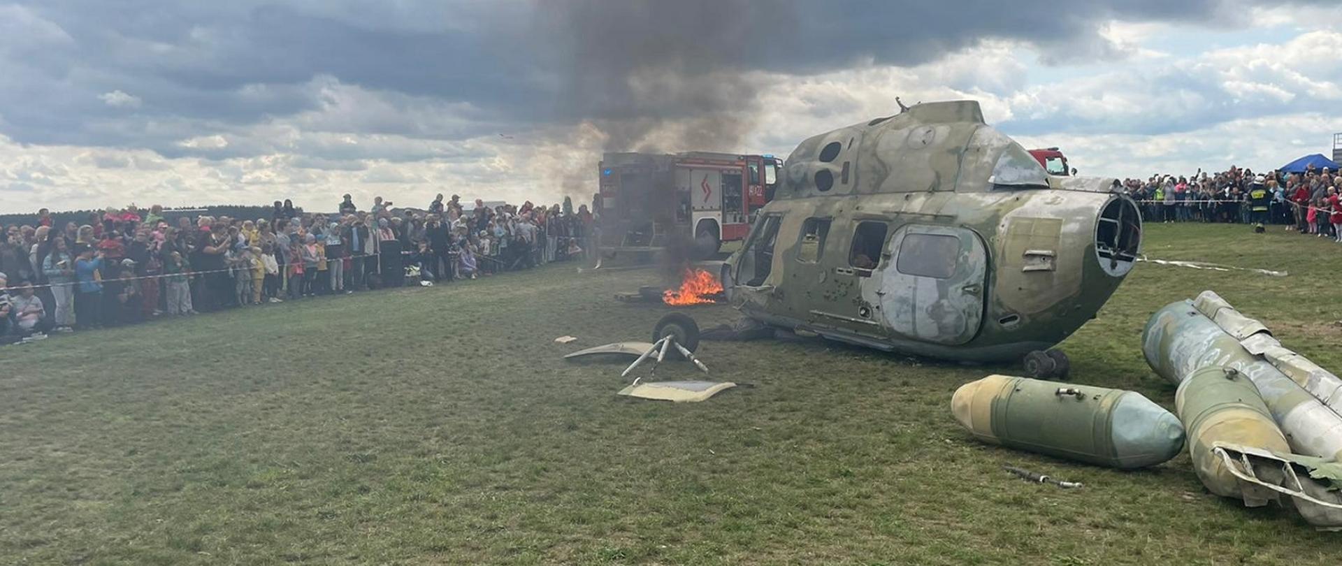 Zdjęcie obrazuje wrak śmigłowca, pożar, samochód strażacki oraz publiczność.