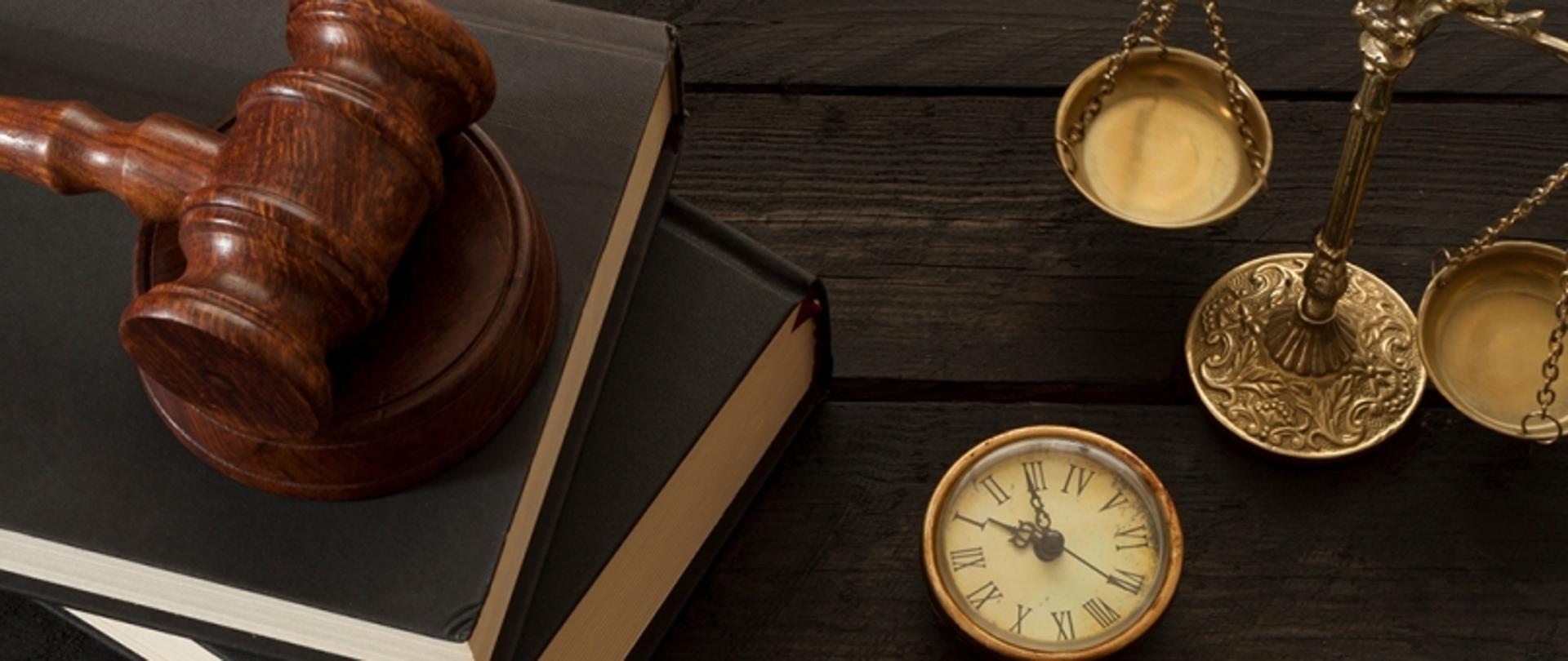 Symboliczne przedstawienie wymiaru sprawiedliwości i porządku prawnego – drewniany młotek sędziowski, waga oraz zegarek kieszonkowy na tle ciemnego drewna, nawiązujące do formalnych aspektów procesu budowlanego