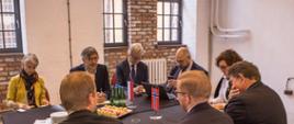 Dwa połączone czarne stoły, na nich flagi Norwegii oraz Polski. Wokół siedzący, elegancko ubrani mężczyźni oraz jedna kobieta prowadzący rozmowy.
