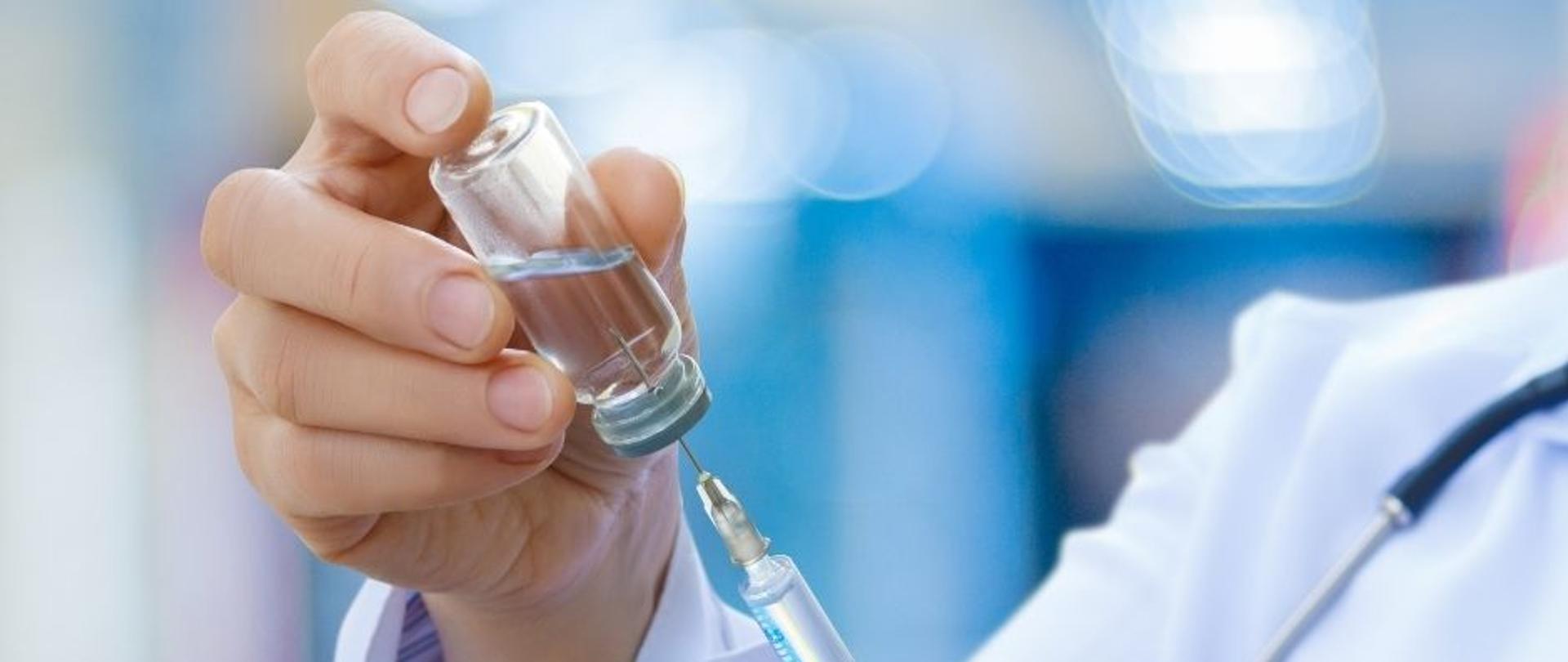 Lekarz pobiera strzykawką szczepionkę z fiolki. Na zdjęciu widoczne dłonie, fragment białego fartucha oraz stetoskop.