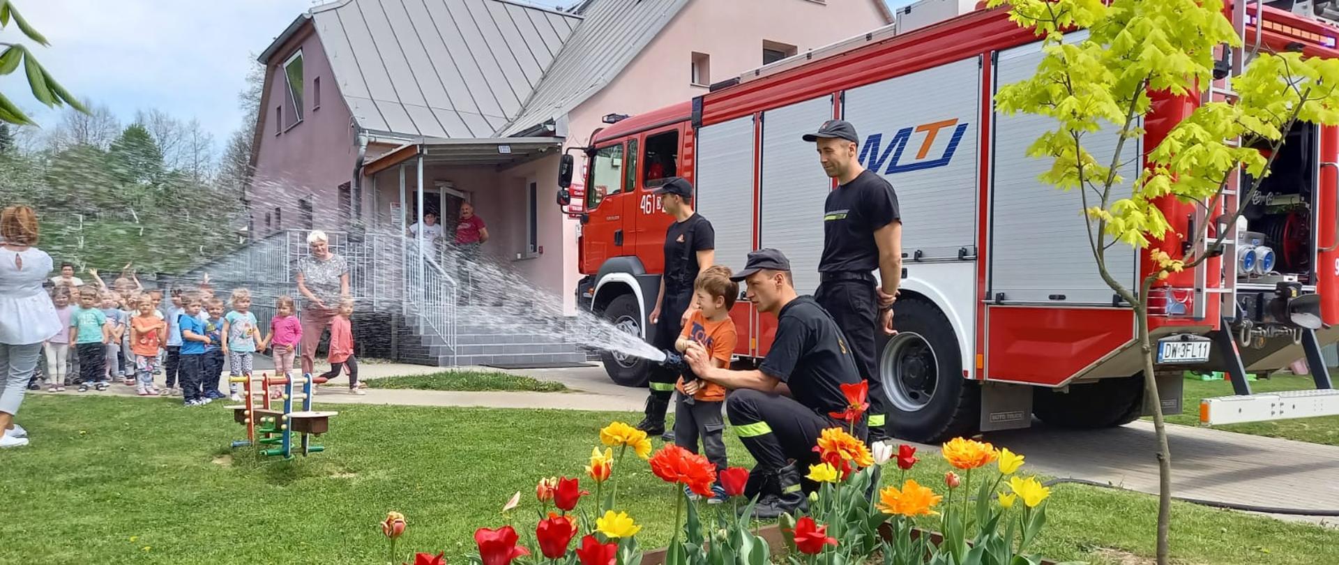 Plac przed budynkiem przedszkola. Po prawej stronie stoi samochód ratowniczo-gaśniczy. Przy nim 3 strażaków w umuindurowaniu koszarowym. Jeden z nich przykląkł przy przedszkolaku i pomaga w utrzymaniu prądownicy wodnej. Po lewej stronie w tle stoi grupa przedszkolaków wraz z opiekunkami.