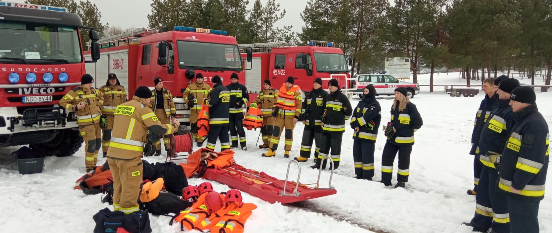 Zdjęcie przedstawia funkcjonariuszy Jednostki Ratowniczo–Gaśniczej Komendy Powiatowej PSP
w Gołdapi oraz strażaków Wojskowej Straży Pożarnej i Ochotniczej Straży Pożarnych w Dunajku (na tle pojazdów pożarniczych), podczas wstępu teoretycznego, w tym zasad BHP (prowadzone przez odpowiednio wyszkolonego strażaka) realizowanego przed przystąpieniem do ćwiczeń praktycznych. Na obrazie widoczny są również sprzęt, który będzie wykorzystywany podczas ćwiczeń, m.in.: ubrania do pracy w wodzie, kamizelki asekuracyjne, kaski ochronne, sanie lodowe i liny ratownicze