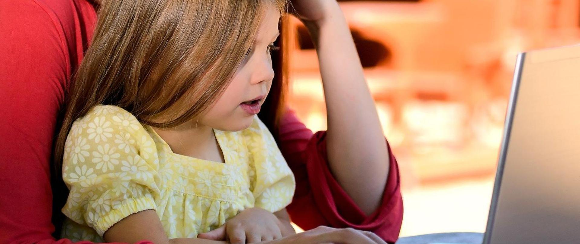 Na zdjęciu mała dziewczynka w żółtym ubranku, która siedzi na kolanach u mamy. Razem wpatrują się w monitor laptopa.