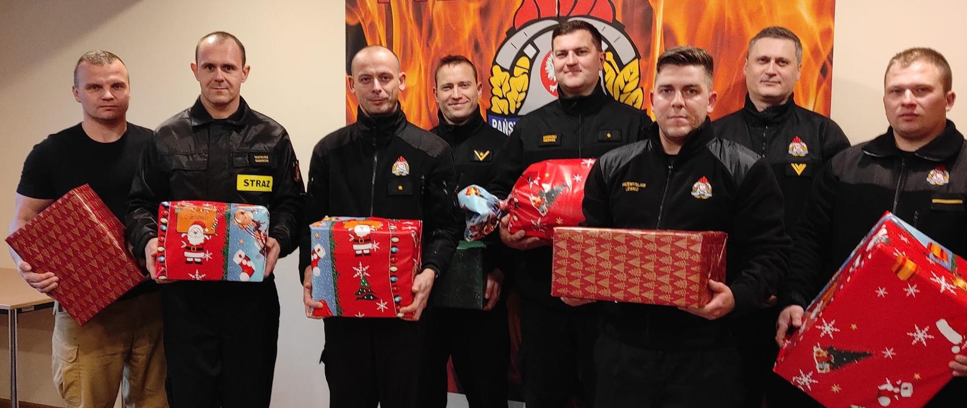 Na świetlicy stoi grupa strażaków w czarnych mundurach w rekach trzymają kolorowe paczki z prezentami.