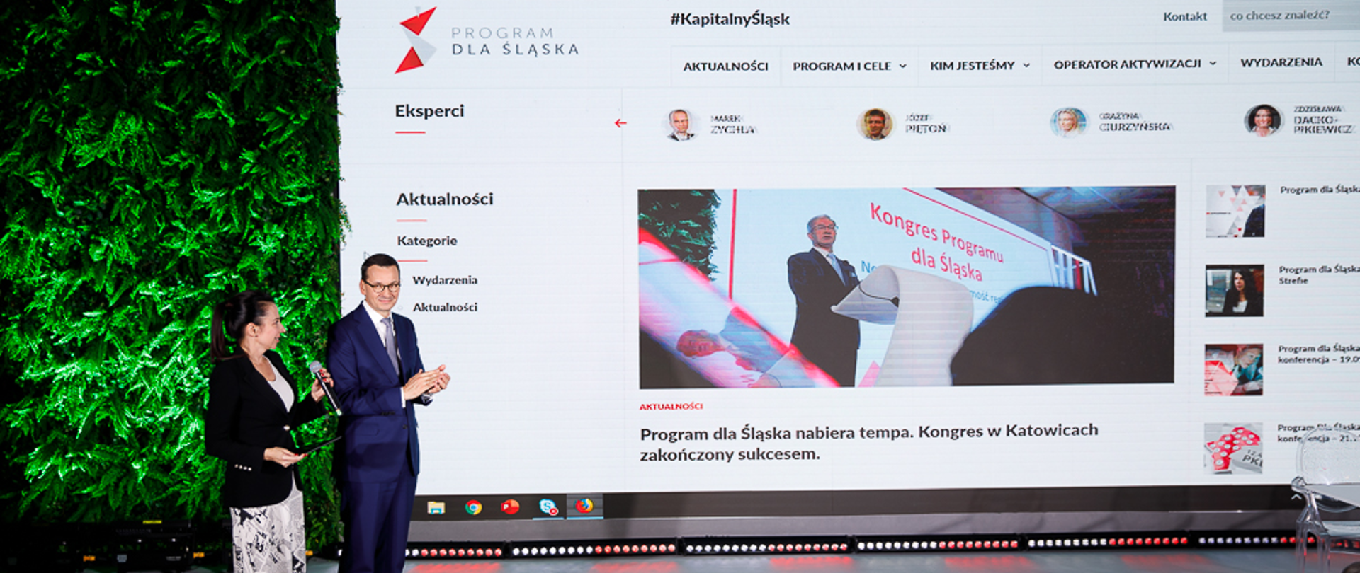 Na zdjęciu dwie stojące osoby - po prawej stronie premier Mateusz Morawiecki, w tle jasny ekran.