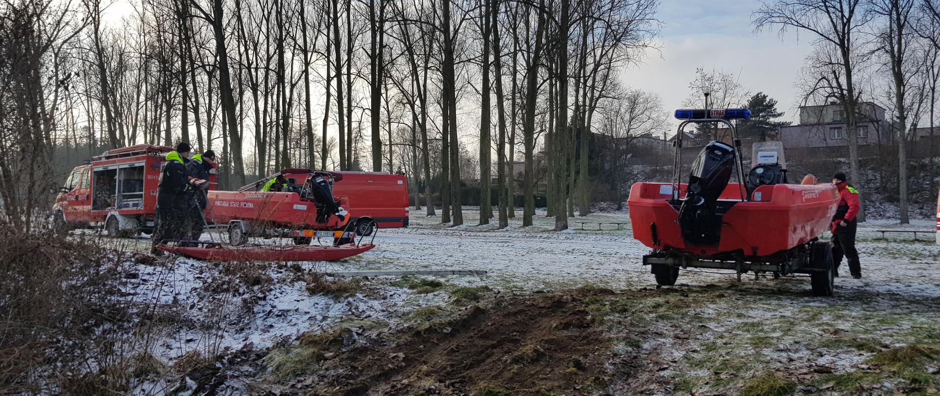 Na zdjęciu widać pojazdy i łodzie straży pożarnej. Pojazdy i łodzie są koloru czerwonego. Miejsce nad rzeką. Ośnieżone. W tle drzewa. Zachmurzenie lekkie. Między pojazdami strażacy.