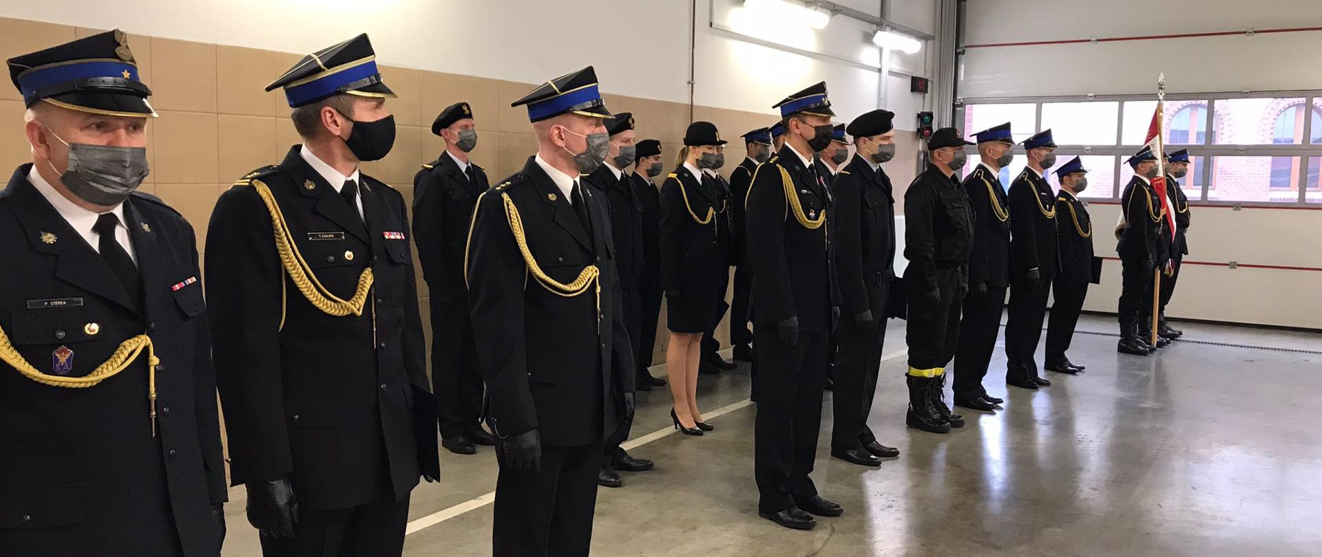 Strażacy w granatowych ubraniach wyjściowych stojący w trzech szeregach podczas zbiórki na garażu JRG1 w Bydgoszczy, na głowach czapki rogatywki, berety, Panie mają kapelusze, wszyscy maski na twarzach, w tle ściany pokryte płytkami ceramicznymi koloru piaskowego do wysokości około 2 metrów, wyżej ściana pomalowana na biało, podłoga szara betonowa. 