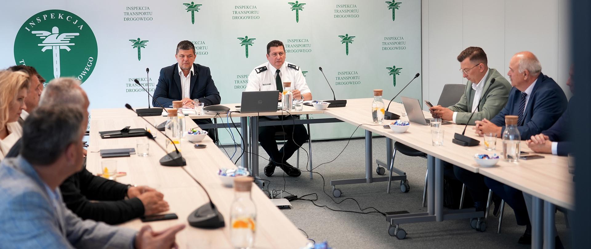 Ogólny widok sali konferencyjnej podczas spotkania przedstawicieli Inspekcji Transportu Drogowego i organizacji zrzeszających przewoźników drogowych. Główny Inspektor Transportu Drogowego Robert Koźlak zabiera głos podczas spotkania. W tle widoczne są elementy identyfikacji wizualnej Inspekcji Transportu Drogowego.