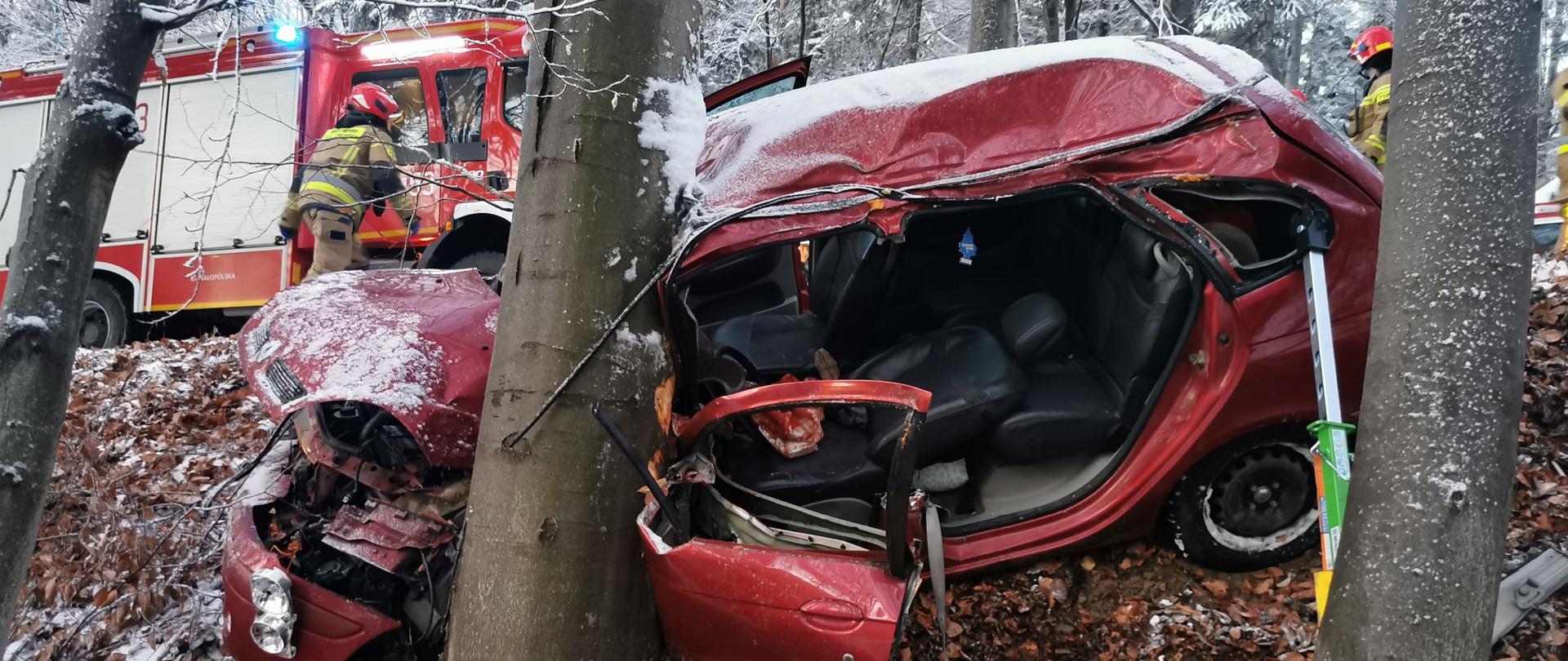 Rozbity samochód osobowy oparty o drzewo. W tle samochód pożarniczy.