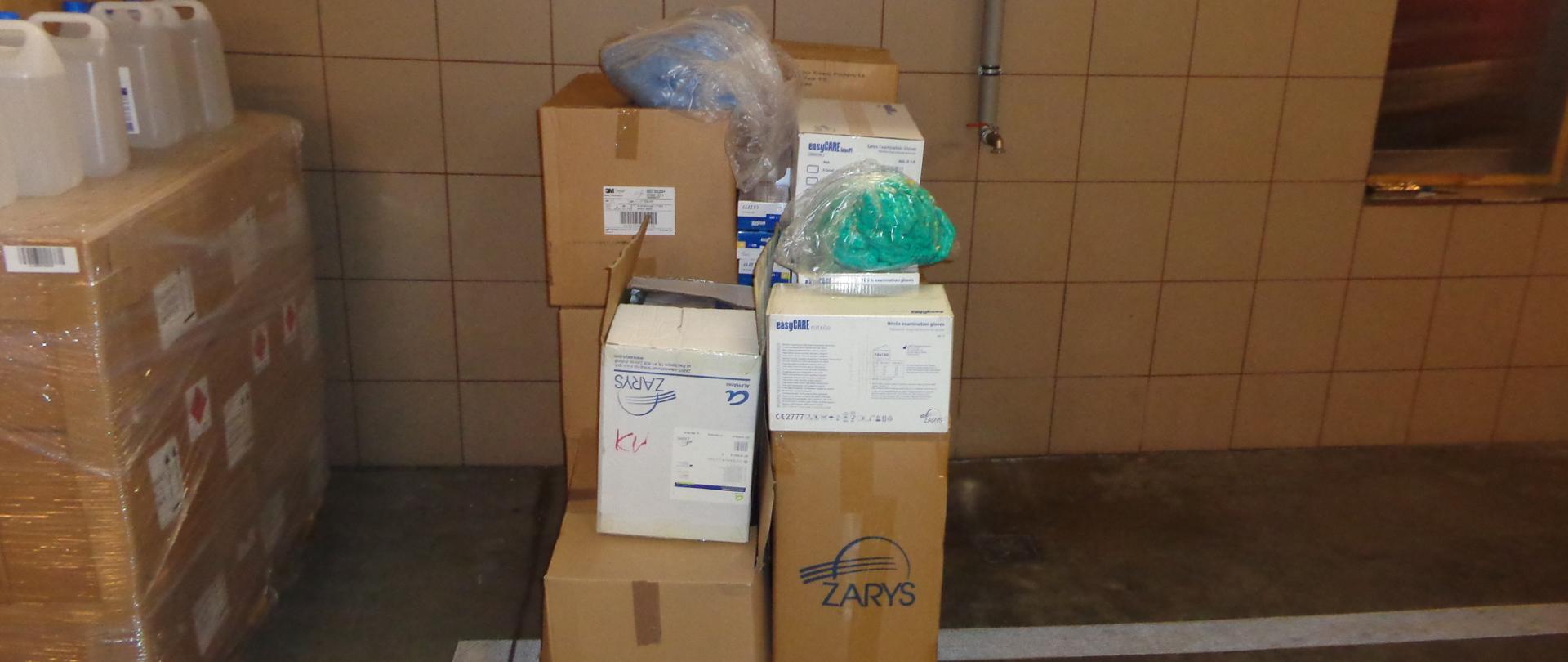 Zdjęcie przedstawia przekazane grodziskim strażakom środki ochrony osobistej znajdujące się w kartonowych pudełkach ułożonych na drewnianych paletach stojących na betonowej posadzce w garażu. W tle ściana wyłożona płytkami w kolorze kawy z mlekiem do wysokości 150 cm, wyżej ścian pomalowana na biało.