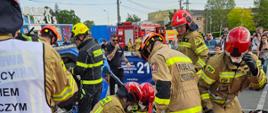 Strażacy udzielają osobie poszkodowanej w symulowanym wypadku pomocy medycznej, w tle działania ratownicze związane z pojazdem, na ziemi sprzęt ratowniczy