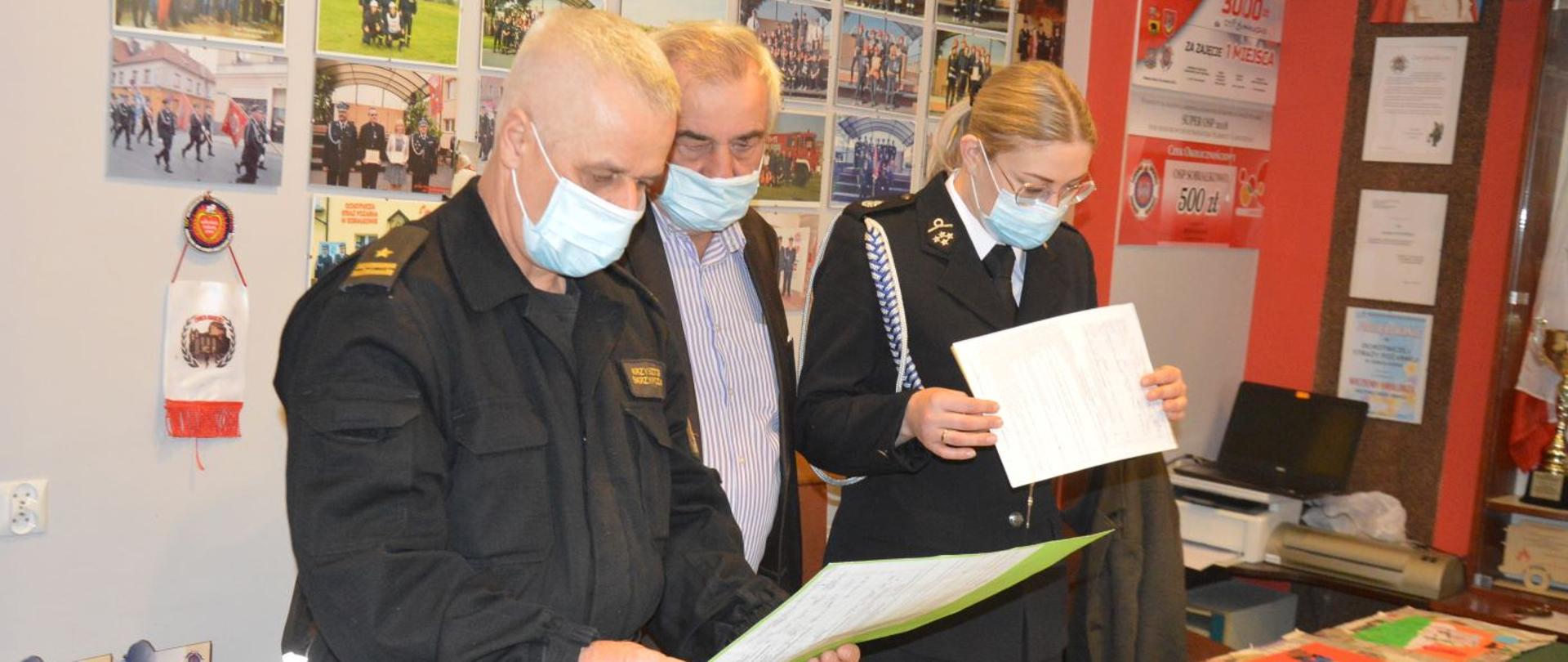 Konkurs plastyczny. Remiza OSP w Sobiałkowie. Jury dokonuje oceny przesłanych prac plastycznych. Strażak PSP oraz druhna OSP przyglądają się trzymanym w rękach pracom. W tle ściana ze zdjęciami. Z prawej strony widoczna jest drukarka.