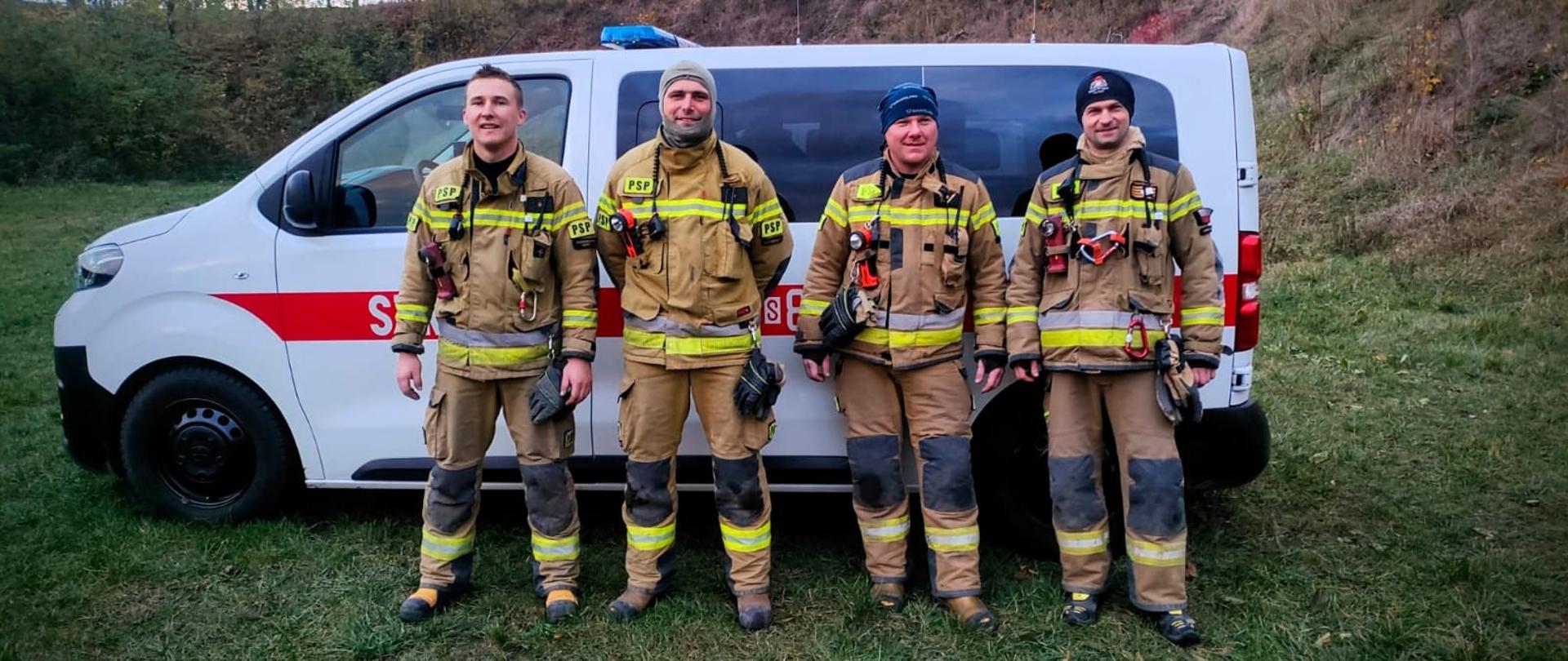 Czterech strażaków w umundurowaniu bojowym stoi na tle białego samochodu strażackiego